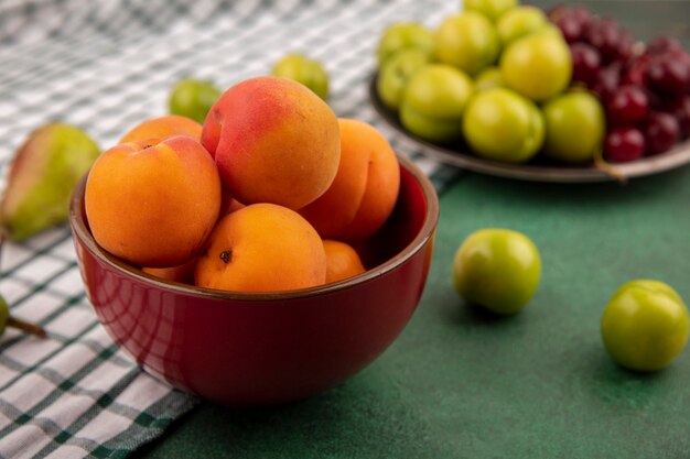 Side view of apricots in bowl and plums cherries in plate with pear on plaid cloth and green background