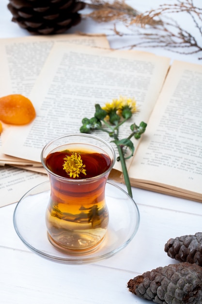 Side view of armudu glass of tea with cinnamon sticks, dandelions and pine cones with an open book on the table