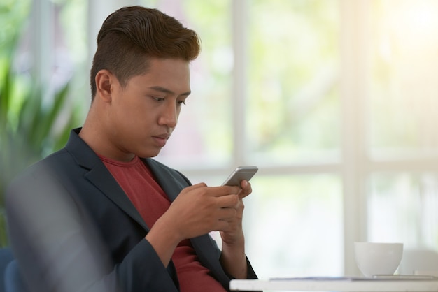 Взгляд со стороны азиатского парня просматривая его smartphone на чашке кофе