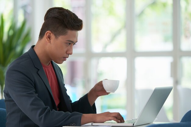 Взгляд со стороны азиатского парня потягивая кофе и работая на портативном компьютере