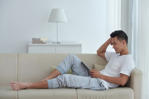 Side view of Asian man seated comfortly on the sofa and watching video on his digital pad