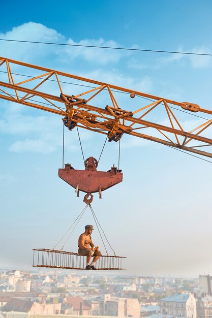 Side view of athletic man in hat sitting and resting on construction on high and eating. Large building crane holding construction with male over city in air. Cityscape and blue sky on background.