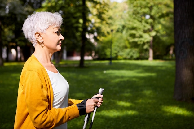 Side view of attractive athletic retired woman with pixie gray haircut enjoying nordic walking using special sticks, doing cardio physical exercise on nice autumn day in countryside. Age and lifestyle