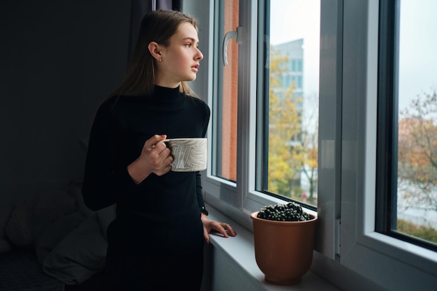 Vista laterale della ragazza attraente che tiene la tazza mentre guarda premurosamente fuori dalla finestra a casa