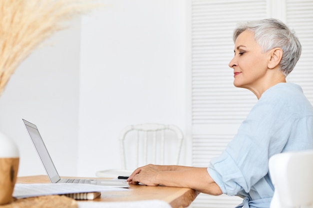 Foto gratuita vista laterale della scrittrice o blogger dai capelli grigi attraente con sguardo pensieroso, seduto davanti al computer portatile aperto, lavorando su un nuovo articolo. senior donna titolare di pensione o di rendita navigare in internet sul dispositivo elettronico