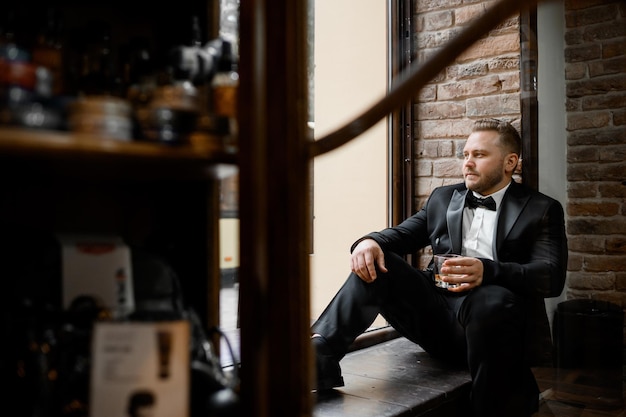 Вид сбоку на привлекательного и вдумчивого жениха, одетого в черный модный костюм и галстук-бабочку, сидящего на лестнице и смотрящего в окно, держа стакан с алкогольным напитком