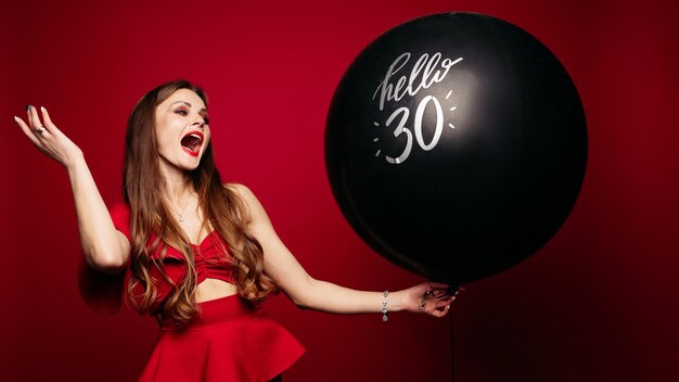 Вид сбоку привлекательной женщины, празднующей день рождения Красивая женщина с макияжем и прической в красном платье держит воздушный шар и смеется на красном изолированном фоне Концепция праздника и счастья