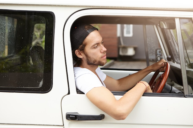 Side view of attractive young bearded hipster sitting on driver's seat while driving his white jeep with his elbow hanging down of open window on sunny day while going to barbecue with his friends