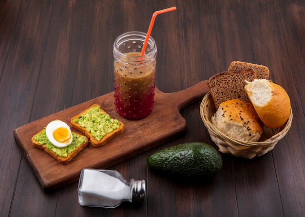 Vista laterale di polpe di avocado su fette di pane tostato con uovo su una tavola di cucina in legno con succo in un bicchiere con un secchio di pane sulla superficie in legno