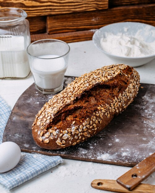 Side view of baked bread with oat flakes and a glass of milk on a wooden cutting board