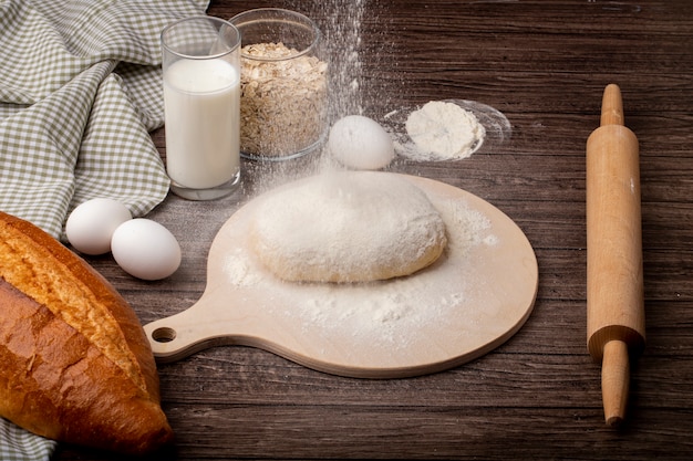 Side view of baking concept with dough and flour on cutting board with rolling pin eggs milk baguette on wooden background