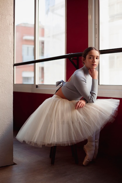Балерина в юбке-пачке позирует рядом с окном, вид сбоку