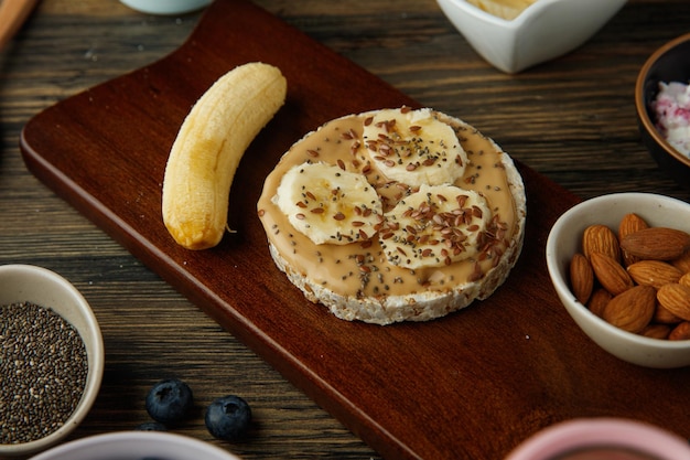 Side view of banana peanut butter cripbread snack with banana and almonds on cutting board and blackthorn on wooden background