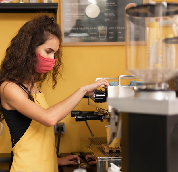 Бесплатное фото Бариста, вид сбоку в медицинской маске во время приготовления кофе в помещении