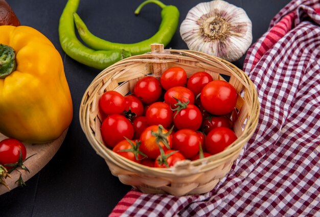 Side view of basket of tomato on plaid cloth with pepper and garlic bulb on black surface