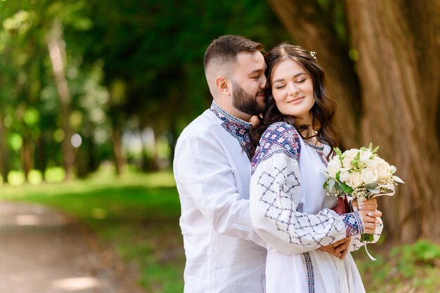 Side view of bearded man which wearing in embroidery shirt standing behide of girl and hugging her waist Attractive bride in long white gown