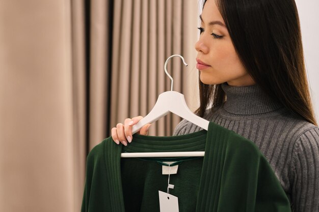 Вид сбоку на красивую азиатскую девушку, задумчиво выбирающую кардиган в магазине современной моды