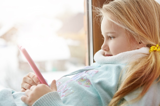 Side view of beautiful blonde little girl sitting at window