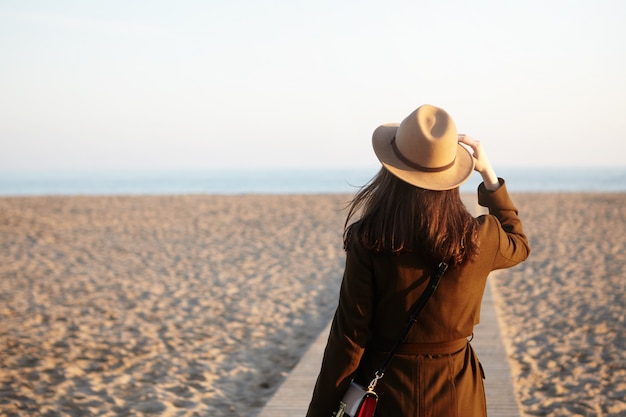 Free photo side view of beautiful female stranger on autumn sand beach. brunette woman looking into distance, noticed ship or dolphin in sea or ocean, adjusting her beige hat with hand, mind full of thoughts