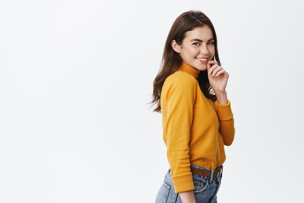 Side view of beautiful girl with white smile touching lip flirty and gazing at camera coquettish standing against white background halfturned