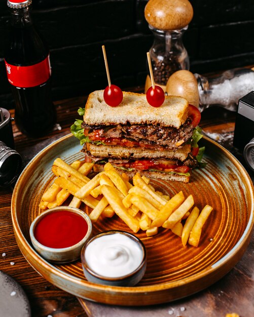 Side view of beef sandwich with tomatoes served with french fries and sauces on plate