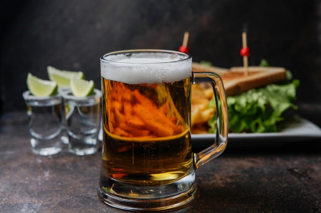 Side view beer with a plate of sandwich and fried potatoes and tequila in a glass served with limes and salt