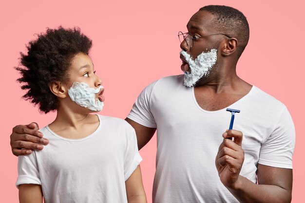 Side view of black father and son stare at each other, have shaving gel on faces, have surprised expressions