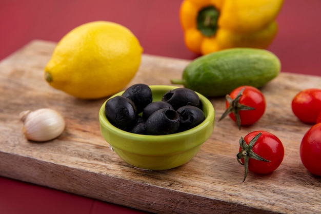 Side view of black and fresh olives in a green bowl on a wooden kitchen board with lemon tomatoes cucumber garlic on red surface