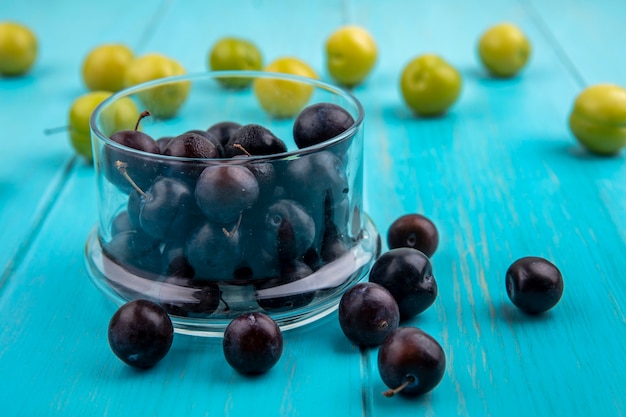 Side view of black grape berries in bowl and pattern of plums and grape berries on blue background