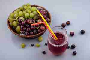 Free photo side view of black grape juice and drinking tubes in glass with grapes in basket and on gray background