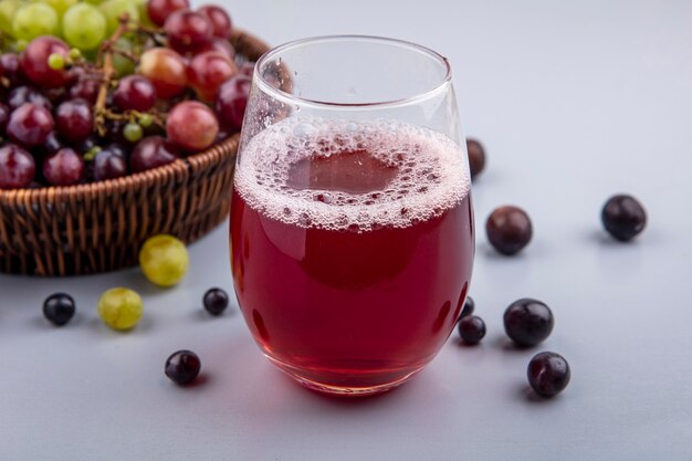 Вид сбоку черного виноградного сока в стакане и корзина винограда с ягодами винограда на сером фоне