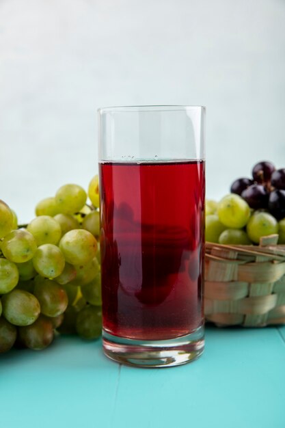 Side view of black grape juice in glass and grapes in basket and on blue surface and white background