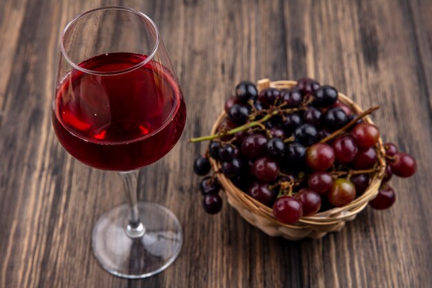 Side view of black grape juice in wineglass with grape in basket on wooden background