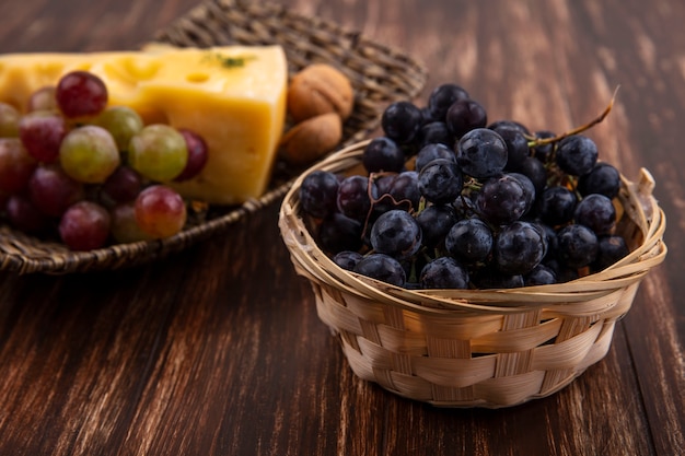 Side view black grapes in a basket with varieties of cheeses and nuts on a stand on a wooden background
