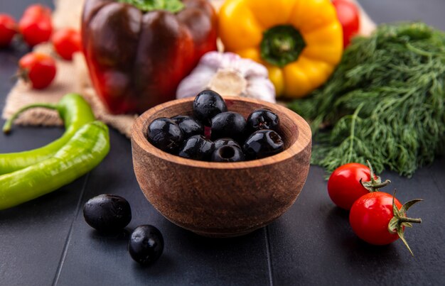 Side view of black olives in bowl with pepper garlic bulb tomato and bunch of dill around on black surface