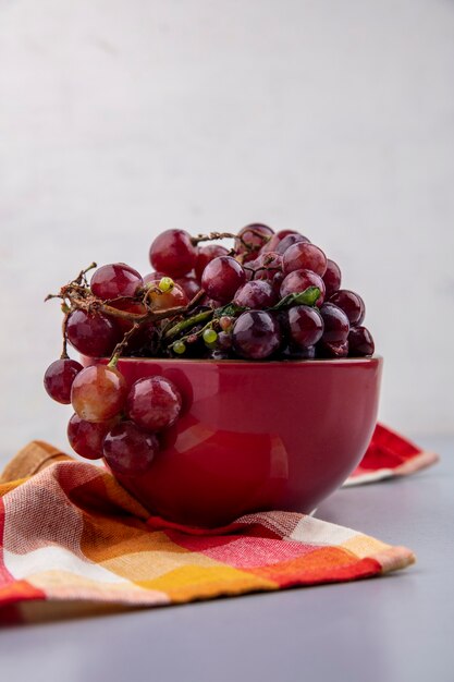 Вид сбоку черного и красного винограда в миске на клетчатой ткани и сером фоне