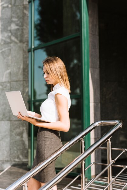 Side view of a blonde young businesswoman standing in front of office looking at laptop