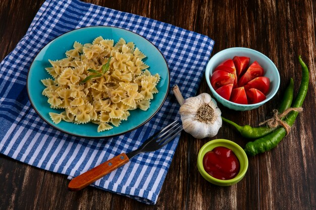 Side view of boiled pasta on a blue plate on a blue checkered towel with a fork tomatoes garlic and chili peppers on a wooden surface