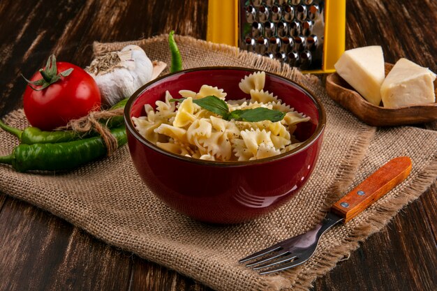 Side view of boiled pasta in a bowl with a fork tomatoes chili peppers garlic and cheese on a beige napkin