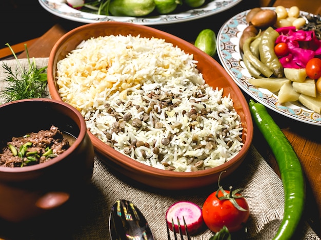 Side view boiled rice on a plate with meat in a pot and salted vegetables