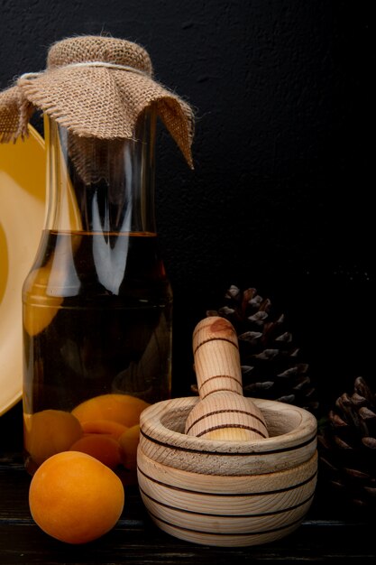 Side view of bottle of apricot compote and garlic crusher with apricot and pinecones on wooden surface and black background