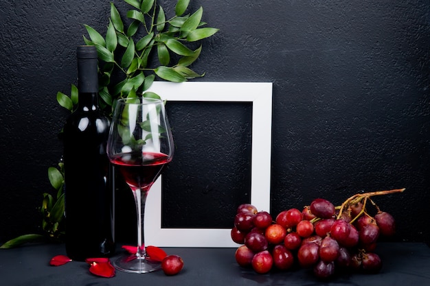 Side view of bottle and glass of red wine with grape frame and leaves with flower petals on black with copy space