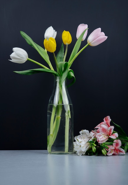 Side view of a bouquet of colorful tulip flowers in a glass bottle and pink alstroemeria flowers lying on the table at black background
