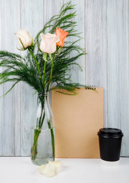 Side view of a bouquet of peach and cream color roses in a glass bottle on the table with a sketchbook and a cup of coffee at grey wooden background