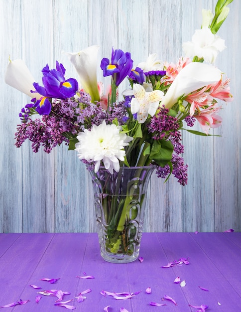 Вид сбоку на букет из белых калл цветных лилий с темно-фиолетовым ирисом, сиреневым белым гладиолусом и розовыми цветами альстромерии в стеклянной вазе на фиолетовой поверхности на сером деревянном фоне