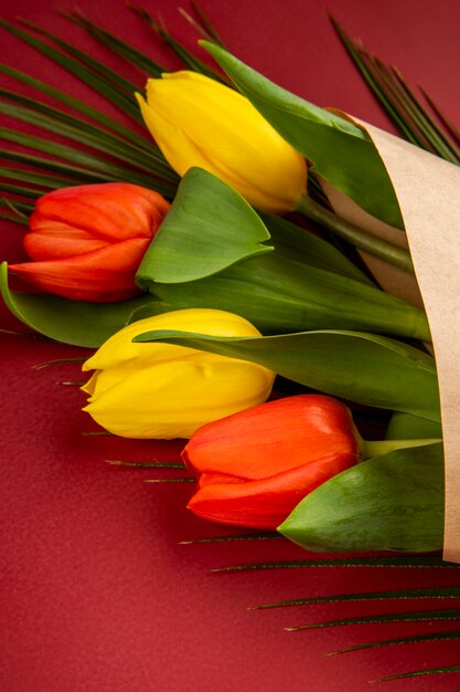 Side view of a bouquet of yellow and red color tulips in craft paper with palm leaf on red table
