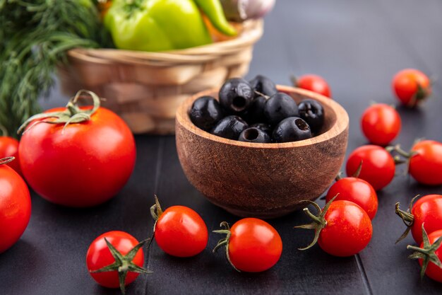 Side view of bowl of black olive with tomatoes around on black surface