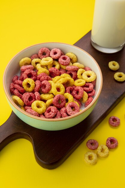 Side view of bowl of cereal with glass of milk on cutting board on yellow background