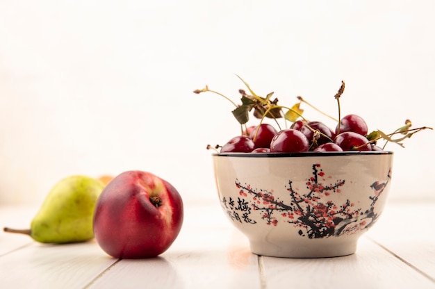 Side view of bowl of cherries with peach and pear on wooden surface and white background