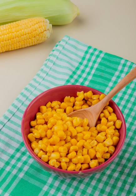 Вид сбоку миску семян кукурузы с ложкой на ткани и початков кукурузы на белом столе